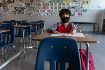 Young boy in mask sitting alone in a classroom at a desk