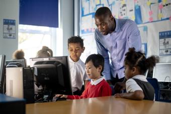 [image of teacher standing behind students who are gathered around a computer]